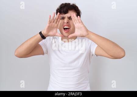 Giovane uomo non binario che indossa una maglietta bianca grida arrabbiato ad alta voce con le mani sopra la bocca Foto Stock