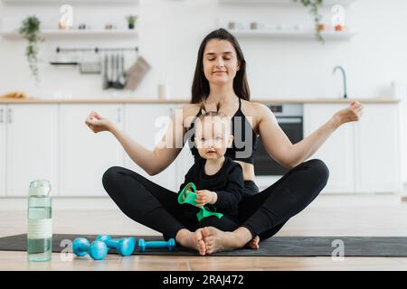 Vista a tutta lunghezza di una donna rilassata seduta in posa yoga con bambino piccolo su un tappeto di gomma in un appartamento moderno. Allenati con le dita nella mudra di gyan mentre la simpatica figlia prova i manubri. Foto Stock