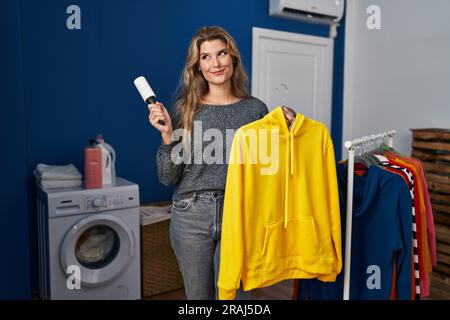 Giovane donna che usa il rotolo di pelucchi per rimuovere i capelli dai vestiti sorridendo guardando lateralmente e fissando via il pensiero. Foto Stock