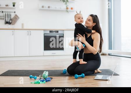 Adorabile ragazza con un vestito nero che si mette in piedi sui giri della donna mentre scambia energia positiva con la mamma durante gli esercizi. Adorabile femmina che abbraccia la figlia con il manichino in mano durante la routine di fitness a casa. Foto Stock