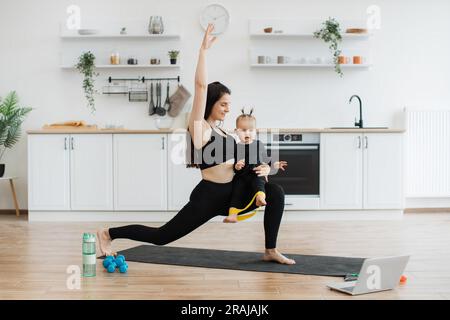 Atleta che indossa abbigliamento sportivo, che fa una passeggiata con il braccio sollevato e un bambino piccolo al ginocchio in una cucina a pianta aperta. Bambino intelligente in abbigliamento sportivo nero che corre in braccio alla mamma con fascia di resistenza sui piedi. Foto Stock