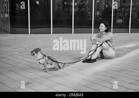 Giovane adulta in città con un cane seduto e abbracciato, sorridente. Accarezzare il cane, nutrirsi, divertirsi Foto di alta qualità Foto Stock