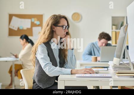 Giovane donna d'affari seduta sul posto di lavoro di fronte al monitor del computer e concentrata sul suo lavoro online Foto Stock