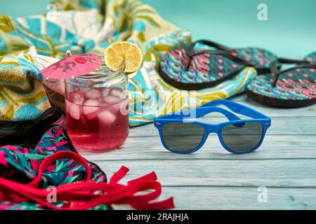 Rinfrescante bevanda estiva con tanto ghiaccio, ombrello e una fetta di limone, occhiali da sole blu, infradito, costume da bagno e telo da spiaggia con motivi tropicali Foto Stock