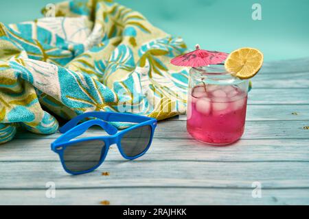 Rinfrescante drink estivo con molto ghiaccio, ombrellone e una fetta di limone, occhiali da sole blu e telo da spiaggia a motivi tropicali su un tavolo di legno e un Foto Stock