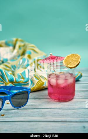 Rinfrescante drink estivo con molto ghiaccio, ombrellone e una fetta di limone, occhiali da sole blu e telo da spiaggia a motivi tropicali su un tavolo di legno e un Foto Stock