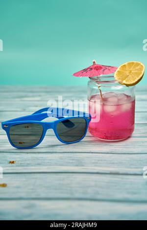 Rinfrescante drink estivo con tanto ghiaccio, ombrello e una fetta di limone e occhiali da sole blu su un tavolo di legno e uno sfondo verdastro. Foto Stock