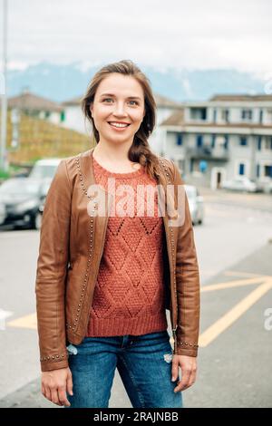 Ritratto alla moda di una giovane donna di 25-30 anni con trucco professionale, con giacca in pelle marrone, pullover a maglia e jeans strappati, p Foto Stock