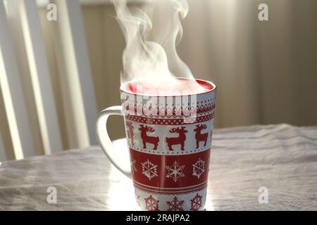 Tazza per la cottura a vapore con caffè o tè sul tavolo. Tovaglia di lino bianco. Primo piano ceramica rosso Natale tazza con fumo sul vecchio tavolo nella sala da pranzo interno bac Foto Stock