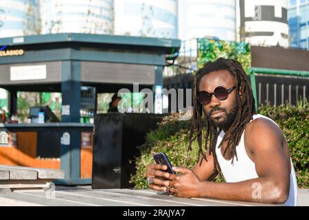 Ritratto di un giovane uomo di etnia africana con dreadlocks e occhiali da sole, seduto alla fiera del cibo all'aperto, in attesa di essere servito dal cameriere, guardando il camer Foto Stock