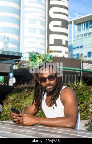 Ritratto verticale di un giovane africano con dreadlock e occhiali da sole, seduto all'aperto in una fiera alimentare all'aperto a Buenos Aires, guardando la macchina fotografica Foto Stock