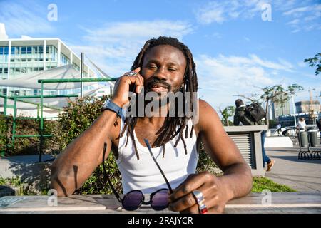 Giovane etnia africana con dreadlock e un atteggiamento cool che tiene gli occhi chiusi felice sorridere parlare al telefono seduto all'aperto in un set urbano Foto Stock