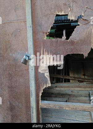 Un piccolo lucchetto attaccato a una porta e a una parete in compensato che è stato danneggiato e presenta dei fori e mostra il contenuto della camera. Foto Stock