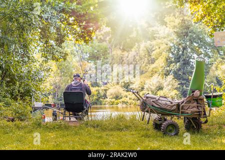 Vista posteriore di un pescatore seduto su una sedia di fronte a una piscina, tenendo in mano una canna da pesca in una mattinata di sole. Attrezzatura da pesca su un carrello in primo piano. Foto Stock