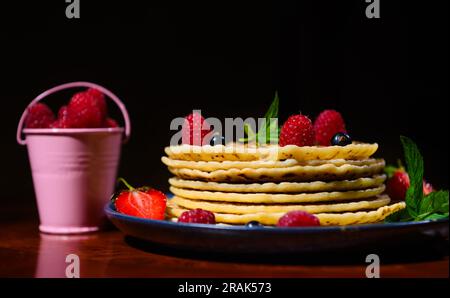 Natura morta. Un piatto di pancake con frutti di bosco freschi e foglie di menta vicino a un secchio con fragole su sfondo scuro. Foto Stock