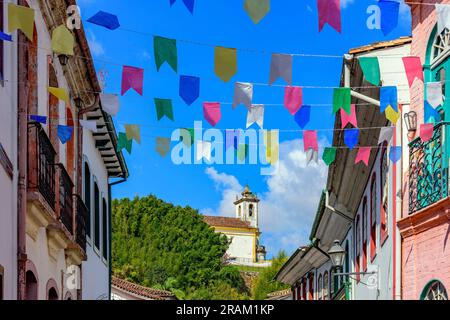 Strada decorata con bandiere per i festeggiamenti di San Giovanni con case in stile coloniale e una chiesa sullo sfondo nella città di Ouro Preto Foto Stock