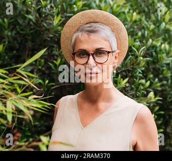 Donna anziana con capelli grigi corti, occhiali e cappello di paglia che guarda la macchina fotografica in un parco Foto Stock