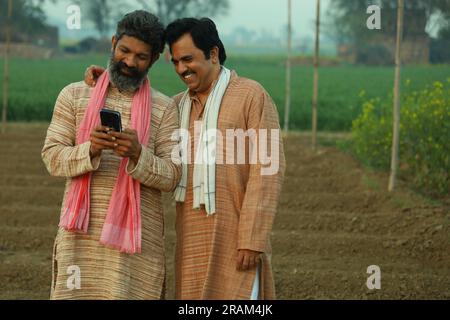 Bella composizione di due contadini indiani in piedi nel campo. Concetto agricolo che ritrae gli agricoltori al lavoro guardando lo smartphone. Foto Stock