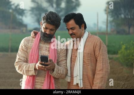 Bella composizione di due contadini indiani in piedi nel campo. Concetto agricolo che ritrae gli agricoltori al lavoro guardando lo smartphone. Foto Stock