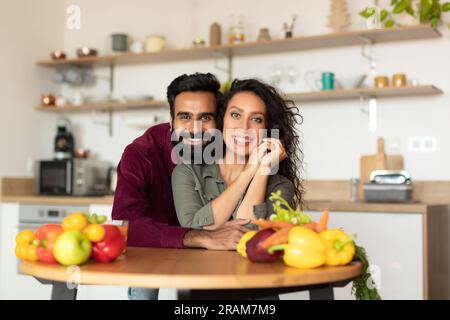 Una coppia araba che si posa insieme in cucina all'interno, un uomo che abbraccia la moglie seduto al tavolo e sorride alla macchina fotografica Foto Stock