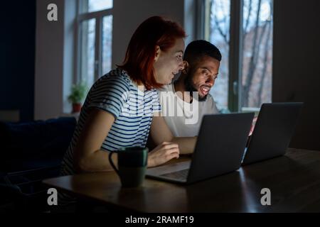 Coppia felice e diversificata ha piccole attività online al lavoro a casa con i laptop la sera Foto Stock
