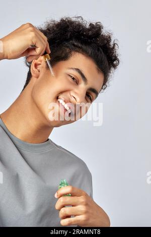 Giovane con i capelli ricci neri idratando la pelle con un olio per il viso in una routine anti-invecchiamento della pelle. Bell'uomo sciarpio che si coccola la pelle Foto Stock