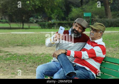 Padre anziano e figlio di mezza età seduti sulla panchina del parco con il telefono cellulare in mano. Videochiama e scatta selfie. Sono immersi in verdi lussureggianti e serenità. Foto Stock