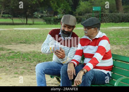 Padre anziano e figlio di mezza età seduti sulla panchina del parco con il telefono cellulare in mano. Videochiama e scatta selfie. Sono immersi in verdi lussureggianti e serenità. Foto Stock