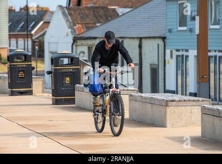 Ciclista maschile che indossa un cappello in bicicletta lungo il lungomare mentre fuma con una sigaretta che gli appende la bocca, nel Regno Unito. Foto Stock