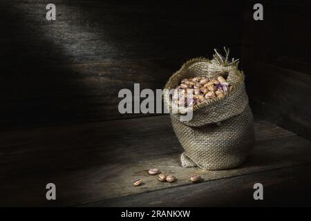 Una natura morta di una pila di fagioli Borlotti crudi, secchi, crudi, crudi, beige, bruni in una borsa assiana nell'angolo di una scatola di legno, in piscine di luce soffusa Foto Stock