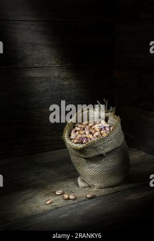 Una natura morta di una pila di fagioli Borlotti crudi, secchi, crudi, crudi, beige, bruni in una borsa assiana nell'angolo di una scatola di legno, in piscine di luce soffusa Foto Stock