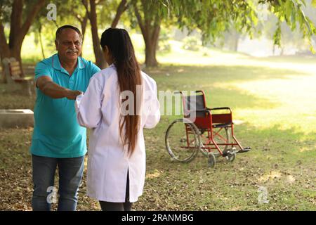 Medico indiano femminile che si prende cura di paziente anziano in giardino dell'ospedale. Il paziente è disabile dal camminare Foto Stock