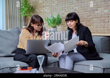 Due donne mature d'affari sedute sul divano in ufficio, a parlare, a parlare di lavoro Foto Stock