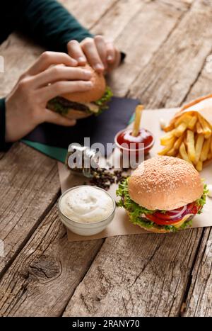 Appetitoso hamburger con succosa cotoletta, patatine fritte, salse su uno sfondo rustico di legno, hamburger in mano sullo sfondo. Concetto di fast food. Foto Stock