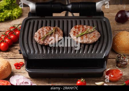 Le polpettine di hamburger di manzo con spezie vengono fritte su una griglia elettrica su uno sfondo rustico di legno. Cucinare hamburger fatti in casa. Ingredienti per hamburger. Foto Stock