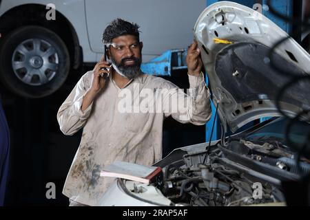 Primo piano del meccanico barbuto Happy Car in piedi nella stazione di servizio accanto a un'auto. Specialista di auto che parla al telefono mentre esamina il motore. Foto Stock