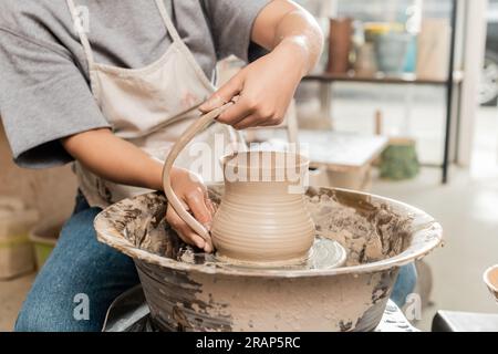 Vista ritagliata della giovane ceramista in grembiule che produce caraffa di argilla mentre lavora con la ruota in ceramica sfocata in un laboratorio di ceramica sullo sfondo, vaso artigianale Foto Stock