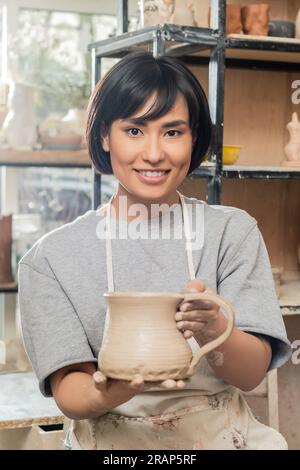 Ritratto di una giovane bruna asiatica artigiana in grembiule e abbigliamento da lavoro che tiene la caraffa di argilla e guarda la macchina fotografica in un laboratorio di ceramica sfocata sullo sfondo Foto Stock