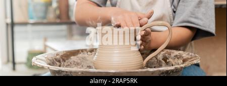 Vista ritagliata di una giovane ceramista sfocata in grembiule e abbigliamento da lavoro che crea una caraffa di argilla mentre lavori con la ruota in ceramica nello studio d'arte sullo sfondo, argilla Foto Stock