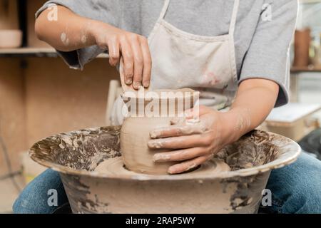 Vista ritagliata di una giovane artigiana in grembiule che rae il vaso di argilla con un utensile in legno mentre si lavora con la ruota di ceramica che gira in un laboratorio di ceramica Foto Stock
