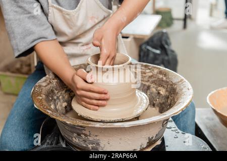 Vista ritagliata della giovane ceramista in vaso di argilla per lo stampaggio del grembiule e lavoro con la ruota di ceramica rotante in un laboratorio di ceramica sfocata sullo sfondo, pott Foto Stock