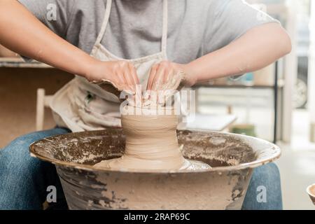 Vista ritagliata della giovane ceramista con lo stampaggio di grembiule in argilla umida e il lavoro con la ruota di ceramica in uno studio di arte sfocata sullo sfondo, scultori di argilla Foto Stock