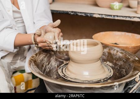 Vista ritagliata della giovane donna della ceramista in abbigliamento da lavoro che tiene l'argilla bagnata vicino alla ruota della ceramica e la ciotola sfocata in uno studio di ceramica sfocata, processo di scultura dell'argilla Foto Stock