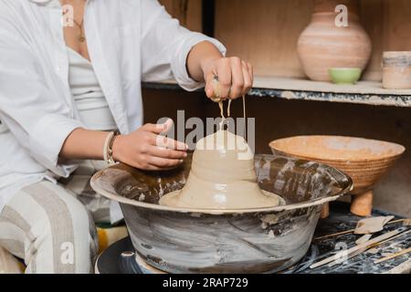 Vista ritagliata di un'artista femminile sfocata in abbigliamento da lavoro che versa acqua sull'argilla sulla ruota in ceramica vicino agli attrezzi e ciotola in un laboratorio di ceramica sullo sfondo, potter Foto Stock