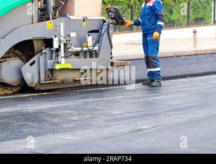 Un frammento del processo di lavoro di un'asfaltatrice sotto il controllo di un ingegnere operatore su una strada cittadina in un giorno d'estate. Copia spazio. Foto Stock