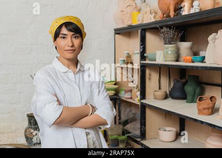 Ritratto di una giovane artigiana asiatica con foulard e abbigliamento da lavoro che incrocia le braccia e guarda la macchina fotografica vicino al rack con sculture in argilla sullo sfondo, vaso Foto Stock