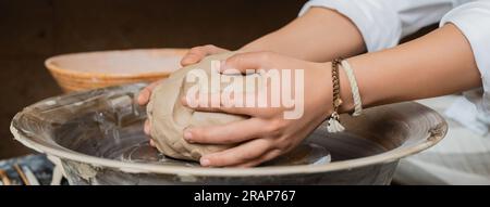 Vista corta della giovane ceramista che mette l'argilla sulla ruota della ceramica mentre lavora vicino alla ciotola in un laboratorio di ceramica sfocata, lavorazione artigianale delle ceramiche a st Foto Stock