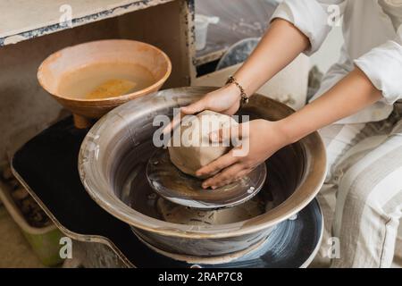 Vista ritagliata di una giovane artigiana donna che lavora in argilla per lo stampaggio e lavora con la ruota in ceramica vicino alla ciotola con acqua in un laboratorio di ceramica, artigianato i Foto Stock