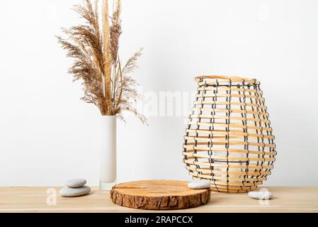 Decorazioni minimali in legno naturale con piedistallo a disco in legno di pino con corteccia, canne secche in vaso e lanterna rotonda in legno a lume di candela, parete bianca. Foto Stock