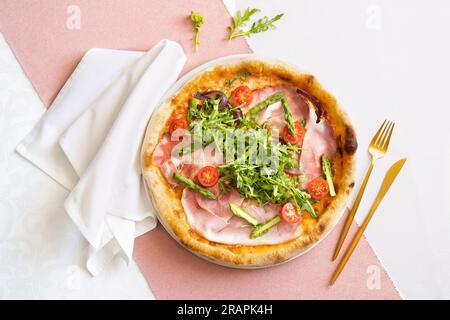 Vista dall'alto di una pizza al prosciutto con prosciutto, asparagi, pomodori e rucola. Tavolo del ristorante con panno rosa e bianco, tovagliolo bianco e posate dorate. Foto Stock